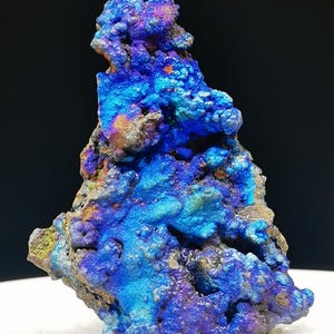 Puede incluir: Un vibrante espécimen mineral multicolor con una superficie rugosa y texturizada. La roca muestra tonos de azul, púrpura y naranja, sobre un fondo oscuro. La forma única y las variaciones de color del mineral lo convierten en una pieza de exhibición llamativa.