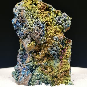 Könnte beinhalten: Ein farbenfrohes, schillerndes Mineralpräparat mit rauer, strukturierter Oberfläche. Der Stein zeigt eine Vielzahl von Farben, darunter Gold, Blau, Grün und Lila. Das Mineral befindet sich auf einer weißen Oberfläche vor einem schwarzen Hintergrund.