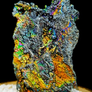 Puede incluir: Primer plano de un mineral gris oscuro iridiscente con una superficie rugosa y texturizada. El mineral presenta una variedad de colores, incluyendo verde, azul, morado y dorado.