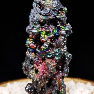 Könnte beinhalten: Ein großes, irisierendes, schwarzes und violettes Mineral mit einer blasigen, strukturierten Oberfläche. Das Mineral liegt auf einem Bett aus weißen Steinen.