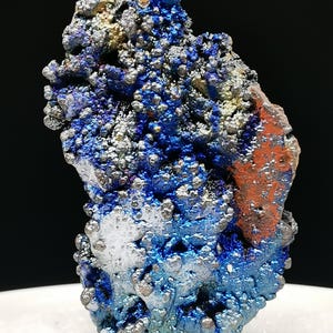 Könnte beinhalten: Nahaufnahme einer Mineralprobe mit strukturierter Oberfläche. Das Mineral weist eine Mischung aus blauen, silbernen und orangen Farbtönen auf. Die blauen Bereiche haben eine blasige Textur, während das Silber metallisch erscheint.