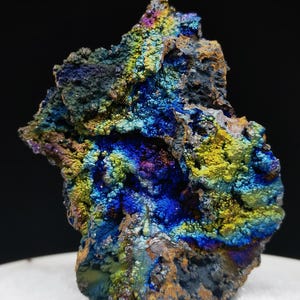 Könnte beinhalten: Nahaufnahme einer farbenfrohen Mineralprobe. Die Gesteinsformation zeigt eine lebendige Farbpalette, darunter Blau-, Gelb-, Grün- und Lilatöne, mit metallischem Glanz. Das Mineral liegt auf einer weißen Oberfläche vor einem schwarzen Hintergrund.