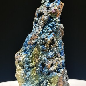Könnte beinhalten: Ein einzigartiges, strukturiertes Mineralexemplar mit einer rauen, unebenen Oberfläche. Der Stein zeigt eine Mischung aus Farben, darunter Blau, Grün und Braun, mit metallischem Glanz. Das Mineral steht aufrecht vor einem schwarzen Hintergrund.