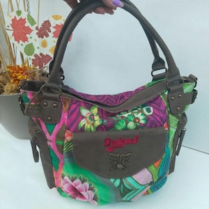 Puede incluir: Un bolso de mano multicolor con un diseño de patchwork con patrones florales y geométricos en tonos verde, rosa y morado. Incluye asas de piel sintética marrón, un bolsillo frontal con la palabra Desigual y un emblema de mariposa de metal.