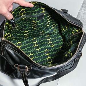 Schwarze Lederhandtasche mit Reißverschluss und weißen Nähten. Das Innere hat ein grün-gelbes Musterfutter. Die Tasche hat einen Schulterriemen und goldfarbene Metallbeschläge.