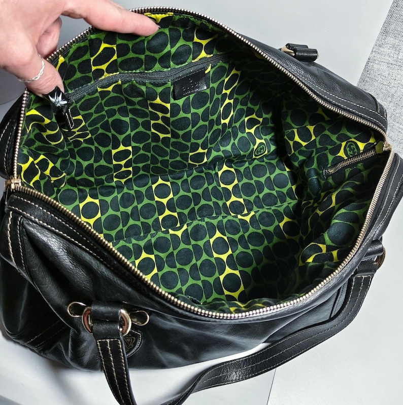 Schwarze Lederhandtasche mit Reißverschluss und weißen Nähten. Das Innere hat ein grün-gelbes Musterfutter. Die Tasche hat einen Schulterriemen und goldfarbene Metallbeschläge.