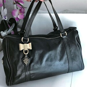 Puede incluir: Bolso de mano de cuero negro con costuras blancas y herrajes dorados. El bolso presenta asas dobles, un adorno en forma de lazo y un charm con el logotipo. Tiene cierre de cremallera y forma rectangular.