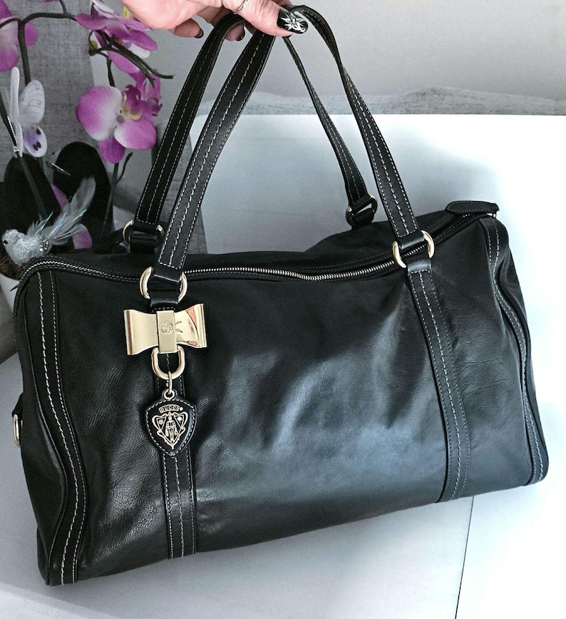 Puede incluir: Bolso de mano de cuero negro con costuras blancas y herrajes dorados. El bolso presenta asas dobles, un adorno en forma de lazo y un charm con el logotipo. Tiene cierre de cremallera y forma rectangular.