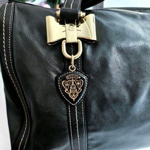 Schwarze Lederhandtasche mit goldfarbener Hardware, darunter ein bogenförmiger Verschluss und ein schildförmiger Anhänger mit dem Markenlogo. Die Tasche hat weiße Nähte und einen oberen Griff.