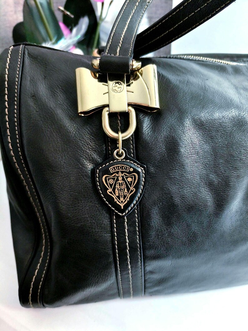 Schwarze Lederhandtasche mit goldfarbener Hardware, darunter ein bogenförmiger Verschluss und ein schildförmiger Anhänger mit dem Markenlogo. Die Tasche hat weiße Nähte und einen oberen Griff.