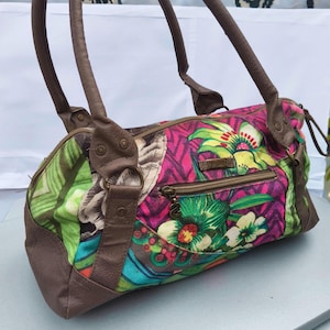 Könnte beinhalten: Eine mehrfarbige Handtasche mit einem lebendigen Blumen- und geometrischen Muster in Pink-, Grün- und Blautönen. Die Tasche verfügt über braune Kunstledergriffe, Besätze und eine vordere Reißverschlusstasche mit einem Metallanhänger.