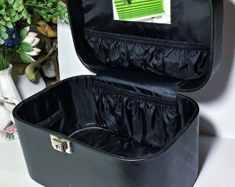 Puede incluir: Maleta de maquillaje vintage negra abierta con un espejo dentro de la tapa. La maleta tiene un cierre y un asa dorados. El interior está forrado con tela negra y tiene un pequeño bolsillo. La maleta es rectangular con esquinas redondeadas.