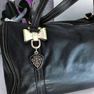 Puede incluir: Bolso de mano de cuero negro con herrajes dorados, incluyendo un cierre en forma de lazo y un charm con el logo. El bolso presenta costuras blancas y asas dobles. El logo de Gucci es visible.