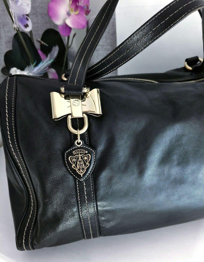 Puede incluir: Bolso de mano de cuero negro con herrajes dorados, incluyendo un cierre en forma de lazo y un charm con el logo. El bolso presenta costuras blancas y asas dobles. El logo de Gucci es visible.