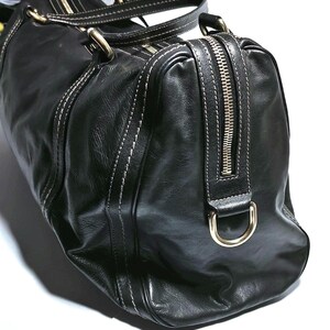 Puede incluir: Bolso de viaje de cuero negro con costuras blancas y herrajes dorados. Cuenta con cierre de cremallera superior y dos asas. El bolso tiene una forma estructurada y un acabado brillante, adecuado para viajes o uso diario.