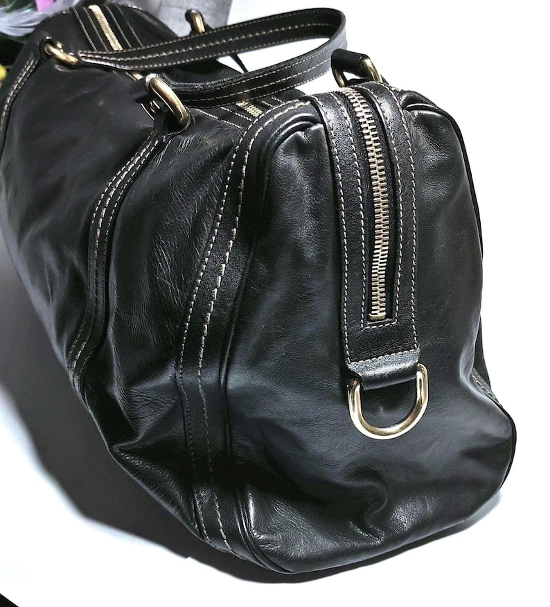 Puede incluir: Bolso de viaje de cuero negro con costuras blancas y herrajes dorados. Cuenta con cierre de cremallera superior y dos asas. El bolso tiene una forma estructurada y un acabado brillante, adecuado para viajes o uso diario.
