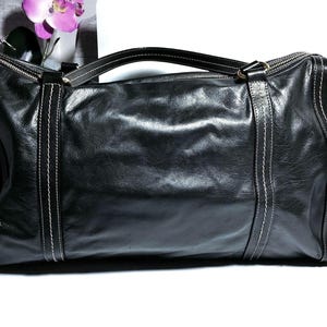 Schwarze Leder-Tasche mit weißen Nahtdetails. Die Tasche hat einen oberen Griff und einen Reißverschluss. Die rechteckige Tasche ist aus weichem Leder gefertigt. Die Tasche ist schwarz und hat ein klassisches Design.