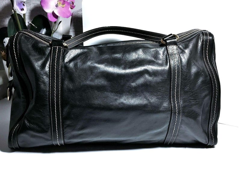 Schwarze Leder-Tasche mit weißen Nahtdetails. Die Tasche hat einen oberen Griff und einen Reißverschluss. Die rechteckige Tasche ist aus weichem Leder gefertigt. Die Tasche ist schwarz und hat ein klassisches Design.