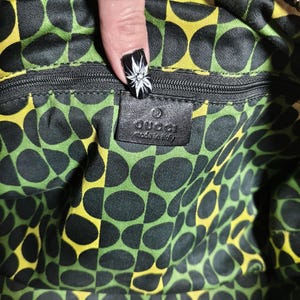 Puede incluir: Interior de un bolso Gucci con cremallera negra y etiqueta de cuero negro. El forro presenta un patr&oacute;n repetido de c&iacute;rculos negros sobre un fondo verde y amarillo. La etiqueta dice "Gucci made in Italy."
