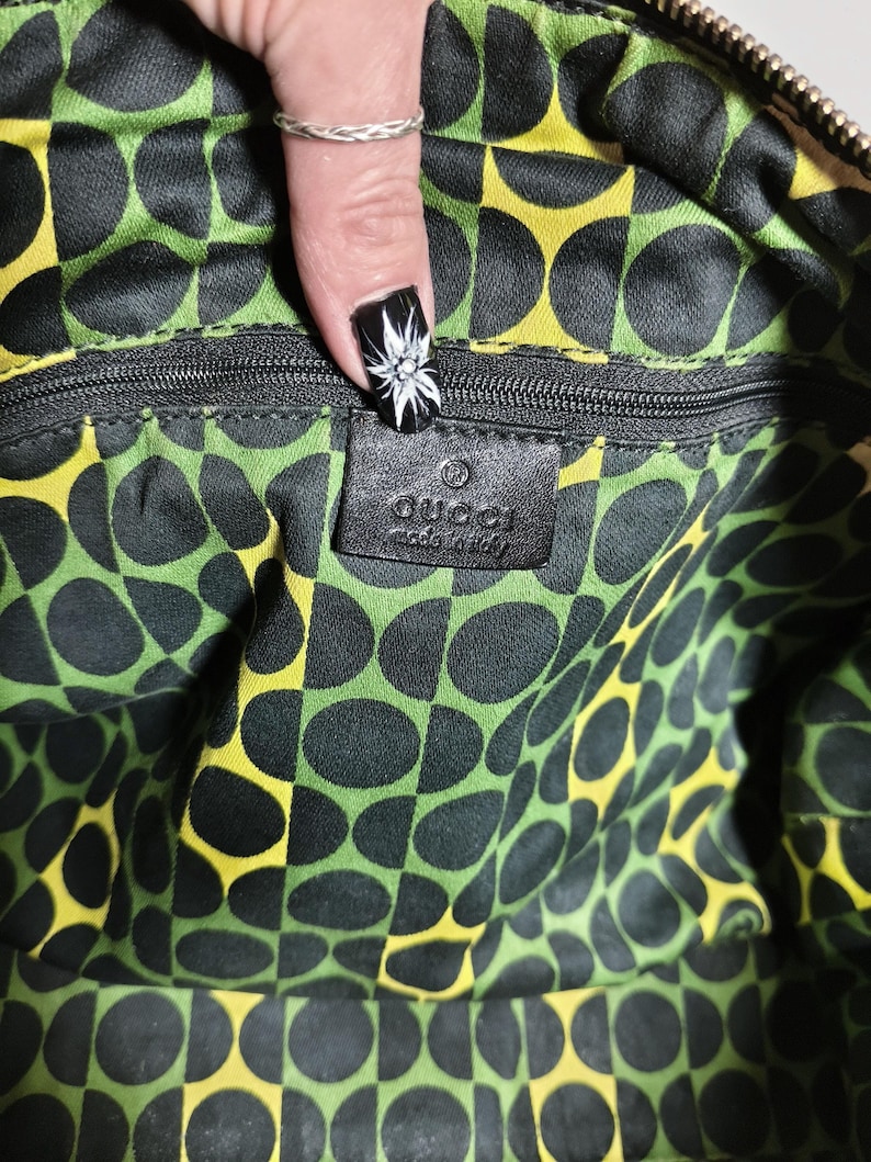 Puede incluir: Interior de un bolso Gucci con cremallera negra y etiqueta de cuero negro. El forro presenta un patr&oacute;n repetido de c&iacute;rculos negros sobre un fondo verde y amarillo. La etiqueta dice "Gucci made in Italy."