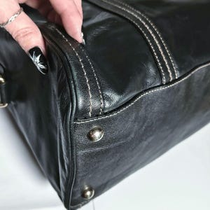 Puede incluir: Primer plano de un bolso de mano de cuero negro. El bolso presenta costuras blancas a lo largo de las costuras y tachuelas de metal plateado en la parte inferior. El cuero parece desgastado, con un aspecto ligeramente envejecido.