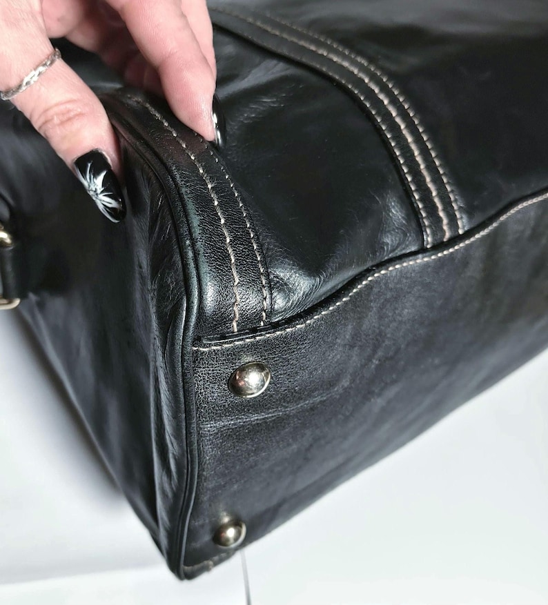 Puede incluir: Primer plano de un bolso de mano de cuero negro. El bolso presenta costuras blancas a lo largo de las costuras y tachuelas de metal plateado en la parte inferior. El cuero parece desgastado, con un aspecto ligeramente envejecido.
