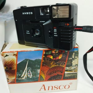 Könnte beinhalten: Schwarze Ansco 35mm Filmkamera mit eingebautem Blitz, auf einer Box mit Reisebildern. Auf der Kamera steht "ANSCO". Das Objektiv ist mit "f5.6 33mm" gekennzeichnet. Die Box zeigt Segelboote und bunte Sonnenschirme. Ein Vintage-Artikel.
