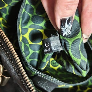 Puede incluir: Primer plano del interior de un bolso de mano de cuero negro con un forro estampado de lunares verdes y amarillos. Una etiqueta con la marca "CI" y el a&ntilde;o "1995" es visible. La cremallera y los herrajes dorados tambi&eacute;n son visibles.