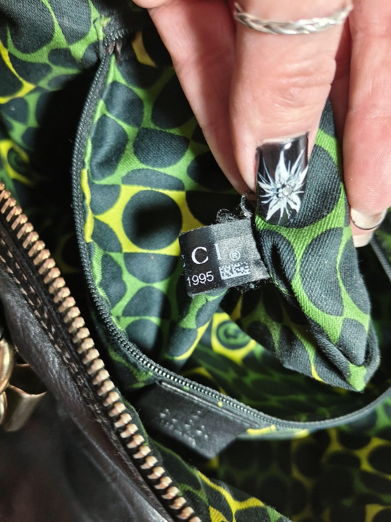 Puede incluir: Primer plano del interior de un bolso de mano de cuero negro con un forro estampado de lunares verdes y amarillos. Una etiqueta con la marca "CI" y el a&ntilde;o "1995" es visible. La cremallera y los herrajes dorados tambi&eacute;n son visibles.