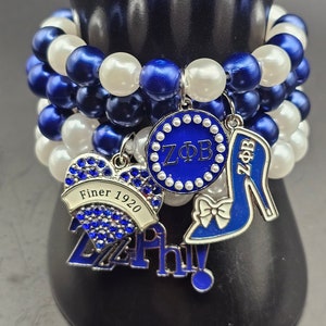May include: A blue and white beaded bracelet with charms. The charms include a heart with the text "Finer 1920", a circle with the text "ZΦB", and a blue high heel shoe with a bow.