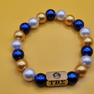 May include: A beaded bracelet featuring navy blue, gold, and white faux pearls. A gold-colored rectangular charm in the center displays the letters "TBS" and the words "TAU BETA SIGMA". The bracelet is set against a yellow background.