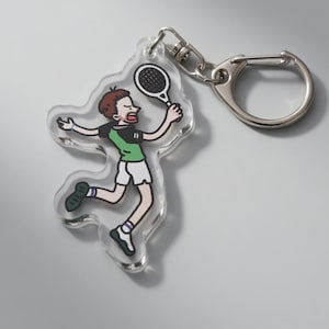 May include: A clear acrylic keychain featuring a cartoon illustration of a person playing tennis. The figure is wearing a green and black shirt, white shorts, and sneakers. The keychain has a silver clasp and ring.