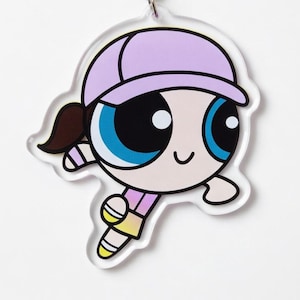 May include: A clear acrylic charm featuring a cartoon character with a light purple cap, brown ponytail, and large blue eyes. The character is wearing a pink and yellow outfit and has a smiling expression. The charm is designed with a white outline.