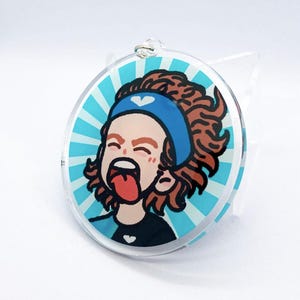Peut inclure: Un porte-clés rond en acrylique bleu et blanc avec une illustration de dessin animé d'une personne aux cheveux bruns, portant un bandeau bleu avec un cœur blanc et une chemise noire avec un cœur blanc. La personne crie la bouche ouverte et la langue tirée.