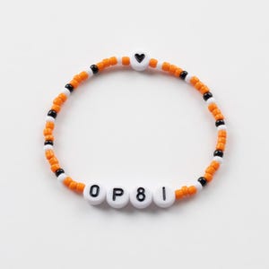 Op de afbeelding: Een armband met oranje, witte en zwarte kralen. De armband heeft witte letterkralen met de tekst "OP81" en een witte hartkralen met een zwart hart. De armband ligt op een witte achtergrond.