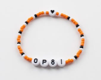 Peut inclure: Un bracelet de perles orange, blanches et noires. Le bracelet comporte des perles de lettres blanches épelant "OP81" et une perle en forme de cœur blanche avec un cœur noir. Le bracelet est sur fond blanc.