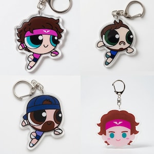 May include: Four clear acrylic keychains featuring cartoon characters with brown hair, each with a silver keyring. The characters have different expressions and wear athletic attire, including headbands and caps. The keychains are on a white background.