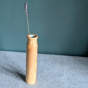 May include: A wooden vase with a light brown finish and a white interior. The vase is tall and slender, with a cylindrical shape. A single dried lavender stem is placed inside the vase.
