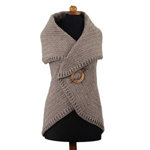 May include: A taupe-colored, crocheted vest with a shawl collar and a wooden pin closure. The vest is displayed on a mannequin and has a textured, handmade appearance. The design features a wrap-style front and a decorative wooden pin.