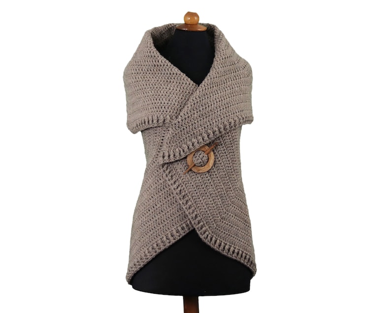 May include: A taupe-colored, crocheted vest with a shawl collar and a wooden pin closure. The vest is displayed on a mannequin and has a textured, handmade appearance. The design features a wrap-style front and a decorative wooden pin.