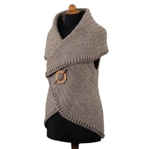 May include: A taupe-colored, crocheted vest with an asymmetrical design. The vest features a wide, shawl-like collar and is secured with a wooden pin. The garment is displayed on a mannequin against a white background.