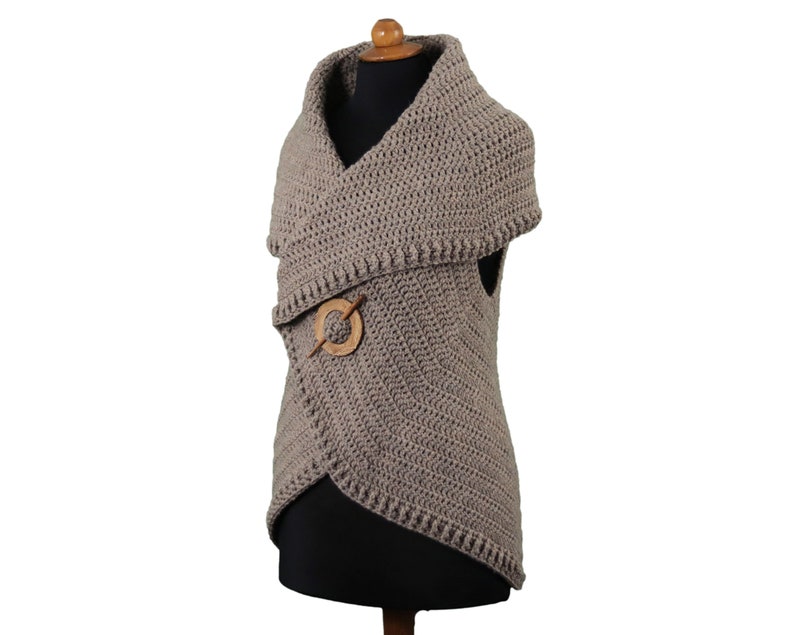 May include: A taupe-colored, crocheted vest with an asymmetrical design. The vest features a wide, shawl-like collar and is secured with a wooden pin. The garment is displayed on a mannequin against a white background.