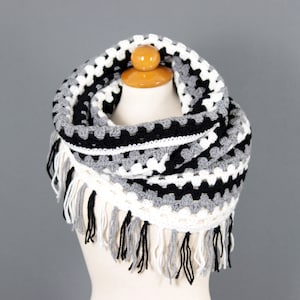 May include: A black, white, and gray crocheted infinity scarf with fringe. The scarf is made with a chunky yarn and has a textured pattern.