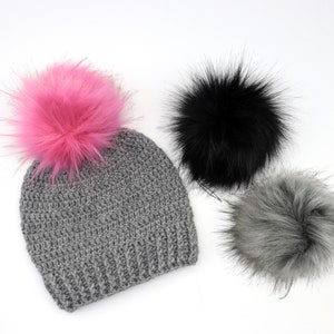 May include: A gray crocheted beanie with a pink faux fur pom pom. There are two additional faux fur pom poms in black and gray.