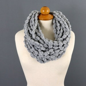 May include: A gray, chunky knit infinity scarf displayed on a mannequin. The scarf is made of a thick, textured yarn, creating a dense, looped pattern. The neutral color and simple design make it a versatile accessory for various outfits.
