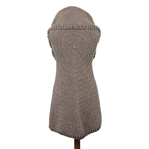 May include: A taupe-colored, sleeveless crochet vest with a hood. The vest features a circular pattern on the back and a textured, open weave design. The garment is displayed on a wooden stand against a white background.