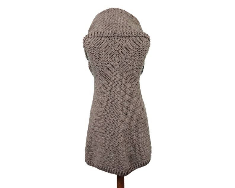 May include: A taupe-colored, sleeveless crochet vest with a hood. The vest features a circular pattern on the back and a textured, open weave design. The garment is displayed on a wooden stand against a white background.