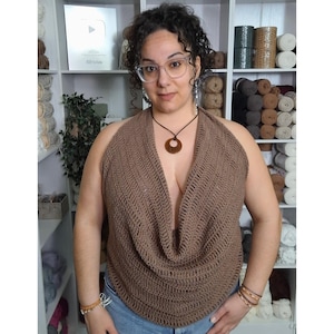 May include: A brown, halter-style crochet top with a deep V-neck. The top is made of a textured yarn and has a draped cowl neckline. The person is wearing a brown pendant necklace and glasses. The background includes shelves with yarn and other craft supplies.