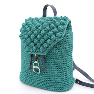May include: Teal crochet backpack with a textured flap and drawstring closure. The bag features navy blue leather straps and silver-tone hardware. The main body of the backpack is a solid teal colour, while the flap has a textured, bobble-like pattern.