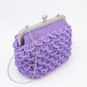 May include: A lavender crochet clutch with a silver metal frame and chain strap. The bag features a textured, scale-like pattern. The clasp is a silver metal ball closure.